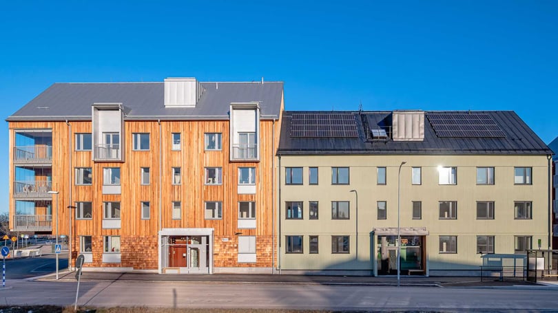 Två flerfamiljshus, ett med träfasad och ett med ljusgrön putsad fasad, stående sida vid sida under en klarblå himmel