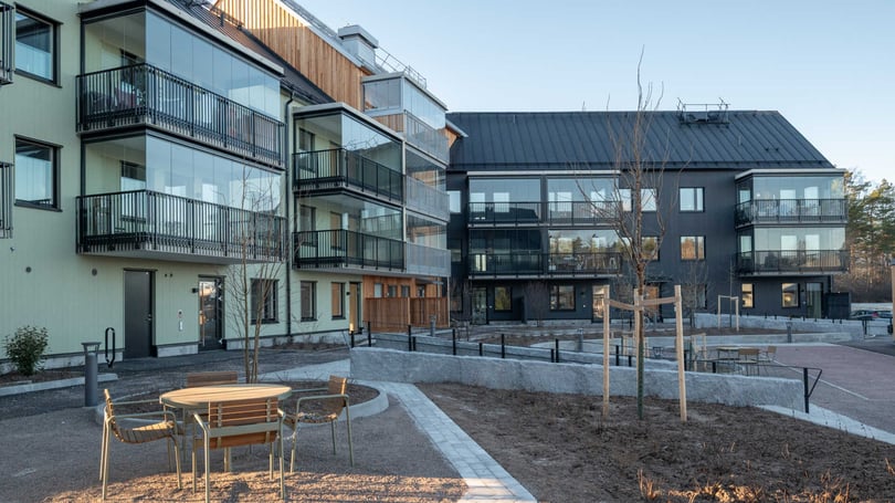 Modernt flerfamiljshus med balkonger, stora fönster och uteplats. Lägenheterna omges av en stenlagd gångväg och ett öppet område med träd