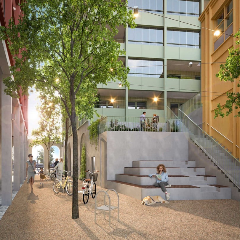 Utomhusmiljö med träd, cykelställ och trappstegsområde vid en modern byggnad. Flera personer sitter och står runt platsen, medan solen skiner mellan byggnaderna