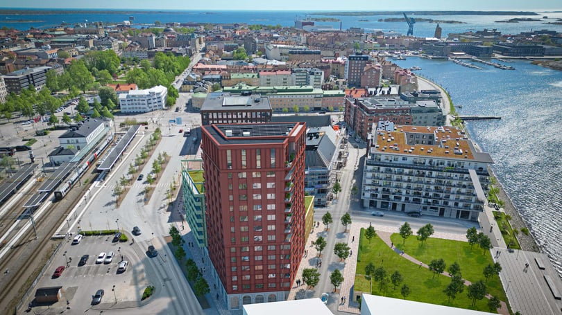 Flygbild över ett stadsområde med en hög byggnad i rött tegel nära en hamn, omgiven av parker, moderna bostäder och en järnvägsstation