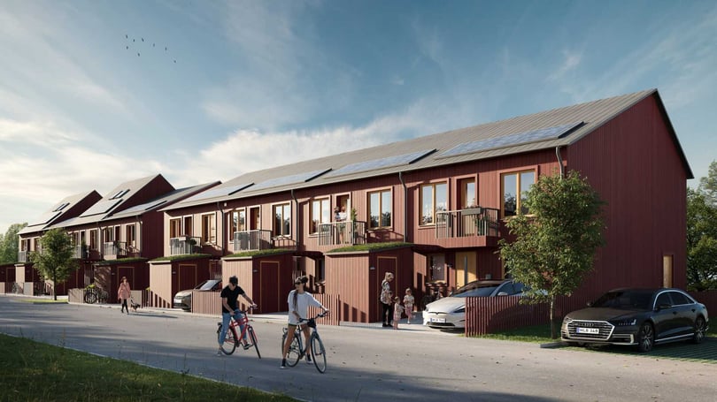 Rad av moderna röda radhus med trädetaljer, solpaneler på taket och omgivande grönska; människor och cyklister på gatan framför husen