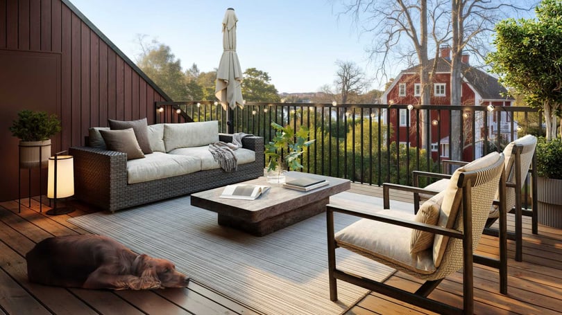 Terrass med trämöbler, soffgrupp, bord, och en liggande hund på ett trägolv, omgiven av natur och röda byggnader i bakgrunden