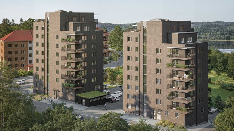 Två moderna flerfamiljshus i brun fasad med balkonger, omgivna av grönska och en väg i ett bostadsområde nära vattnet