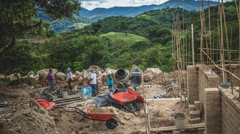 Byggarbetsplats utomhus med flera personer som arbetar, omgiven av natur och berg i bakgrunden