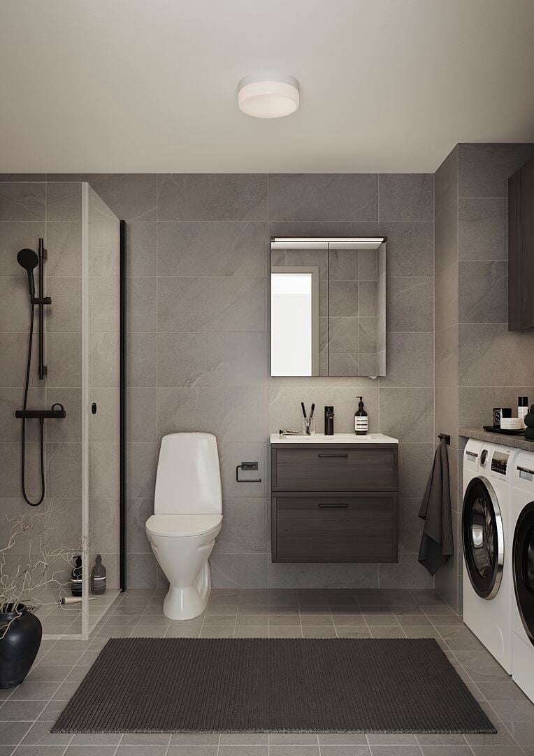Modernt badrum med grå klinkergolv och väggar, vit toalett, dusch med glasvägg, spegel ovanför mörkgrå förvaringsskåp, samt en tvättmaskin och torktumlare