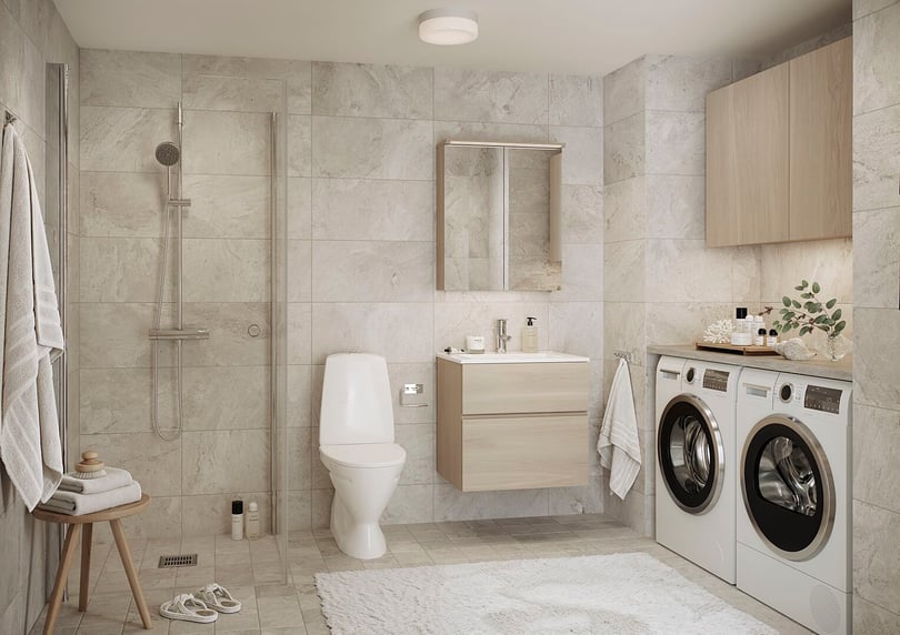 Badrum med ljusgrå kakelväggar, dusch med glasvägg, vita badrumsmöbler, tvättmaskin, torktumlare och badrumsmatta