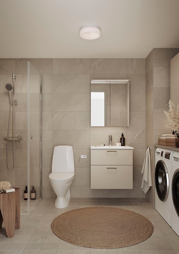 Modernt badrum med duschkabin, toalett, vit kommod med spegel ovanför och två tvättmaskiner bredvid. Neutrala beige väggar och golv med rund jutematta