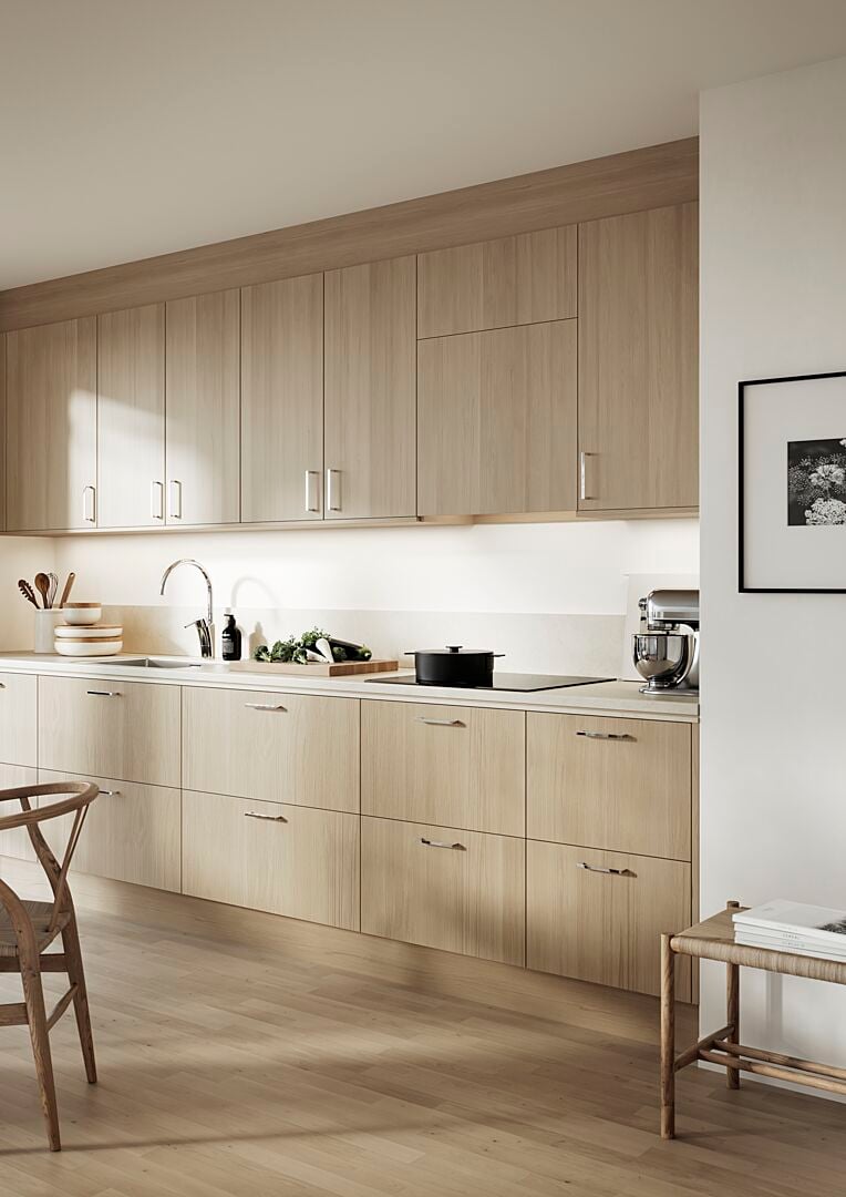 Modernt kök med ljusa träskåp, vit bänkskiva, en diskho med kran och köksredskap. Ett matbord och en stol syns i förgrunden