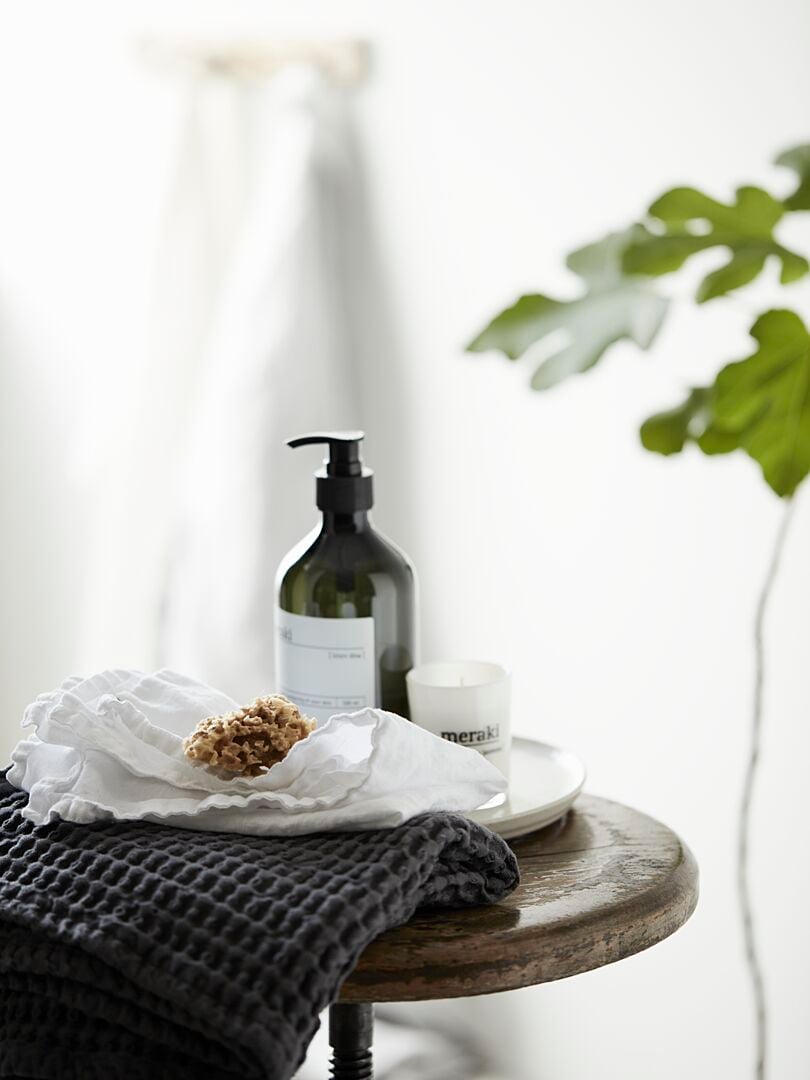Flaskor och burkar med hudvårdsprodukter på ett träbord dekorerat med en handduk och naturlig svamp, i en ljus miljö med en växt i bakgrunden