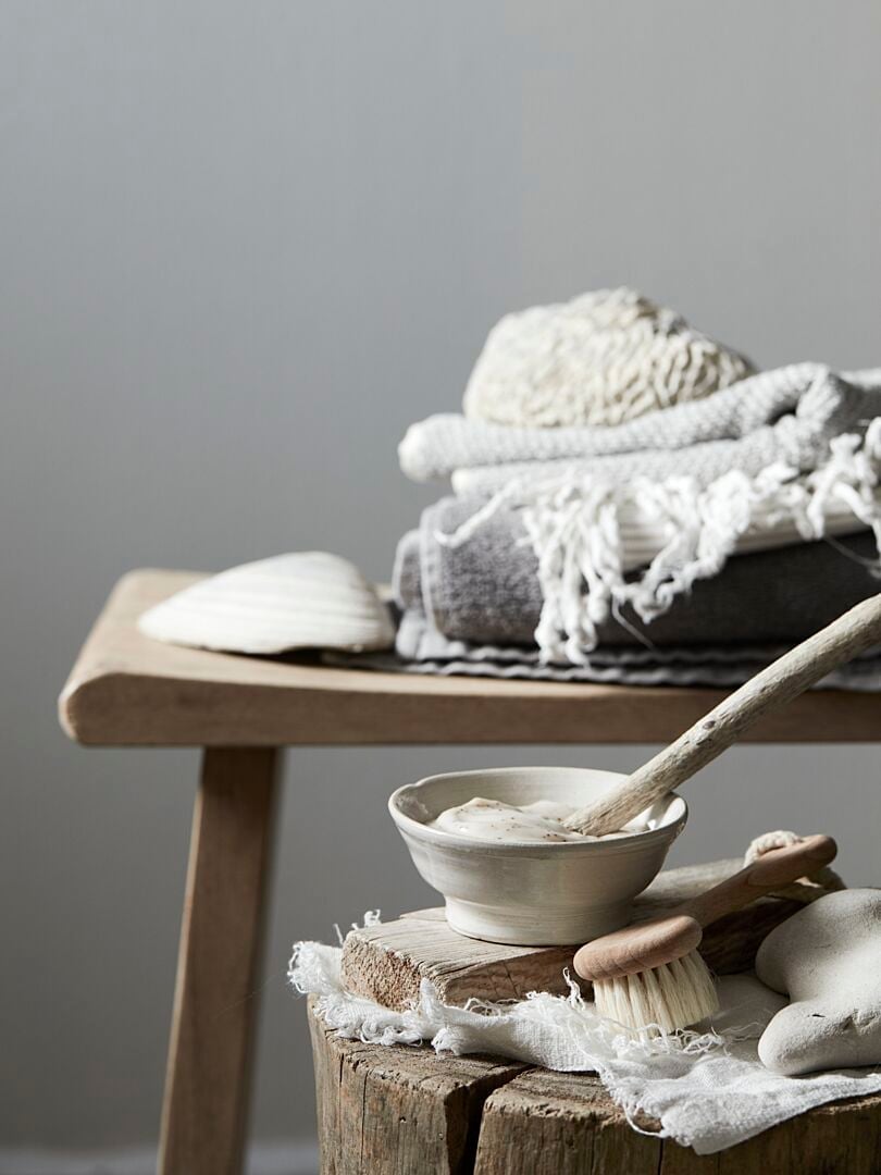 Träbord med vikta textilier, vit skål med kräm och borstar, samt dekorativa stenar i en minimalistisk inredning