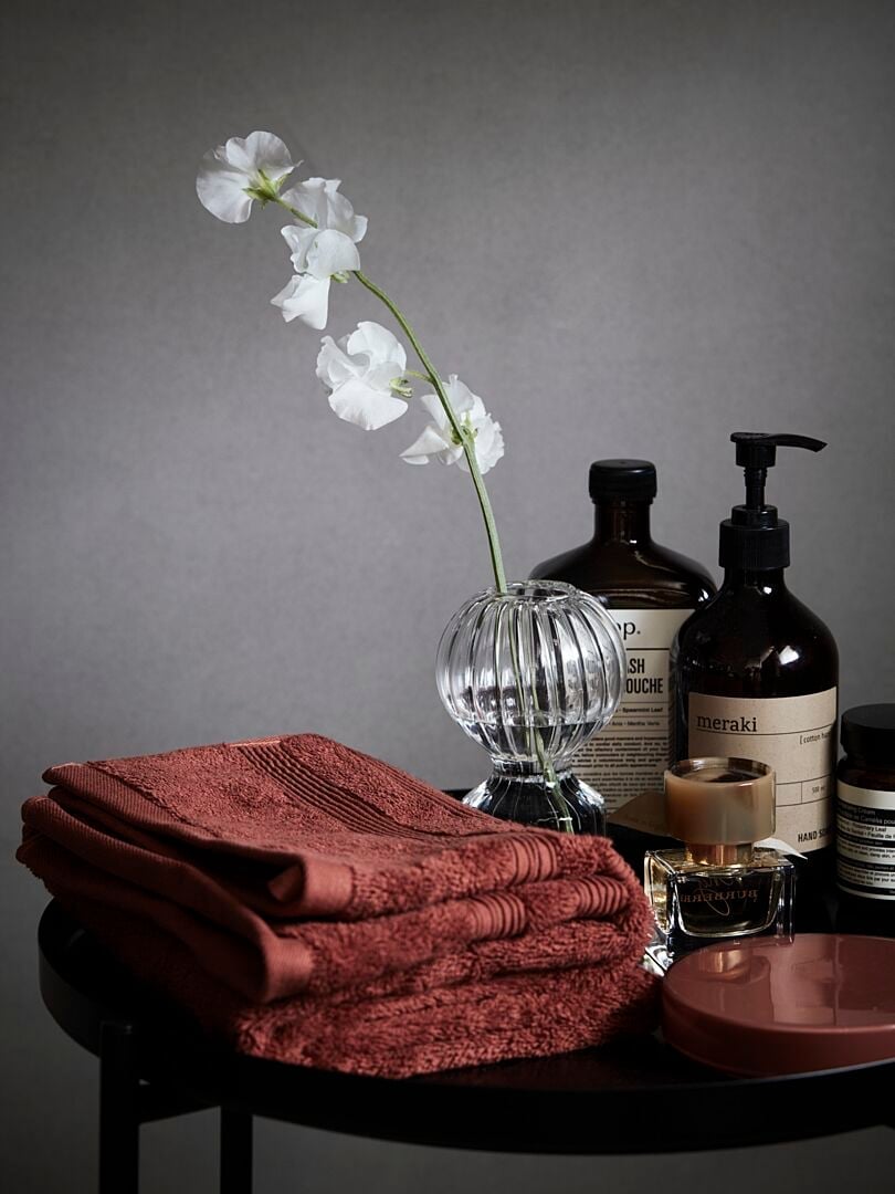Handdukar i roströd färg, glasvas med vita blommor och olika badrumsprodukter på ett svart bord