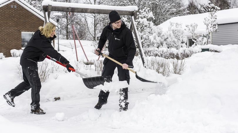 Två personer skottar snö på en snötäckt gård med snöskyfflar; i bakgrunden syns en gungställning, ett hus och snötäckta buskar