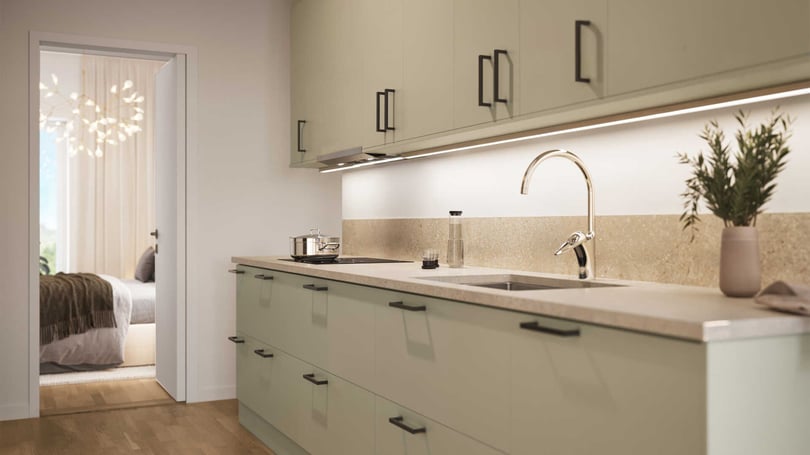 Modernt kök med ljusgröna skåpluckor, svart beslag och bänkskiva i beige ton. En kran, kastrull och vas med grön växt är placerade på bänken