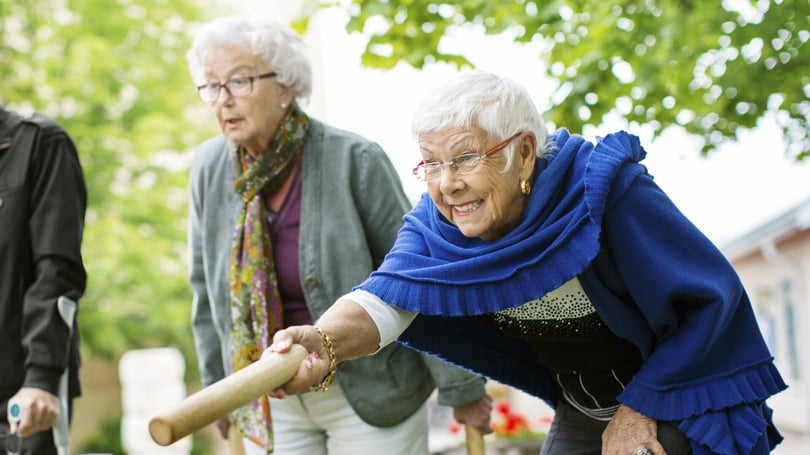 Äldre personer deltar i en utomhusaktivitet, där en person håller i en trästav; bakgrunden består av träd och en byggnad