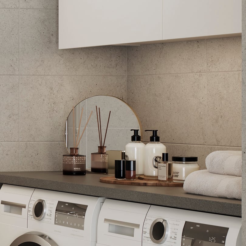 Tvättstuga med två vita tvättmaskiner, handdukar, runda spegeln och dekorativa objekt som tvålpump och doftspridare placerade på en grå bänkskiva