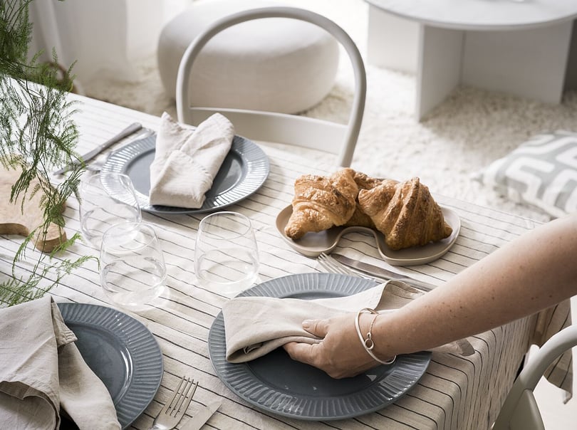 En dukat frukostbord med grå tallrikar, glas, linneservetter och croissanter på ett brunt fat