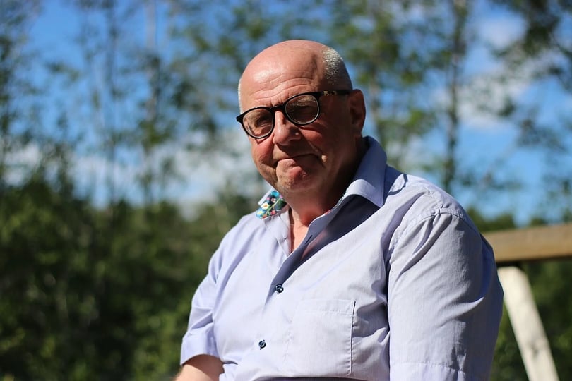 En man i en ljusblå skjorta sitter utomhus med en suddig bakgrund av träd och himmel i dagsljus