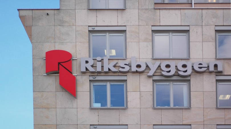 Byggnad med logotypen för Riksbyggen, inkluderande en röd geometrisk symbol och text på fasaden. Bakgrunden visar en klarblå himmel