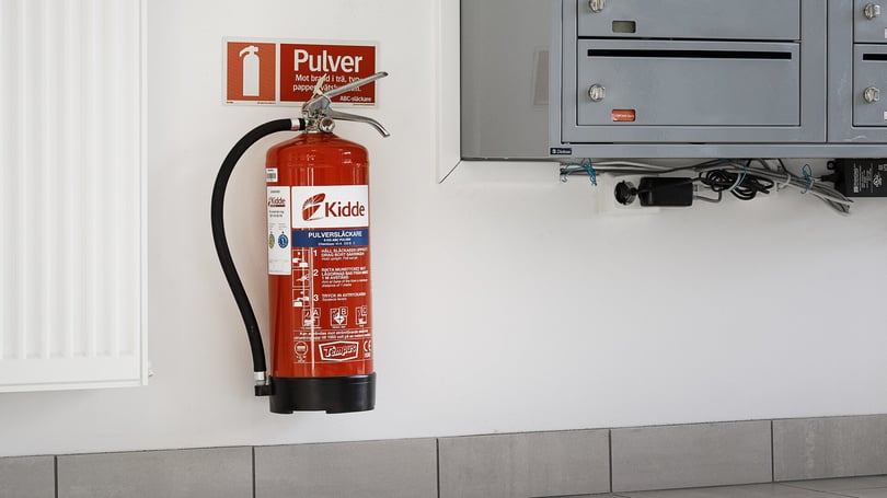 Brandsläckare av typen pulver monterad på en vit vägg bredvid ett grått elskåp