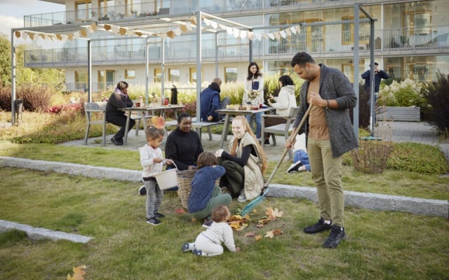 En grupp människor umgås i en utomhusmiljö med gräs och picknickbord; några samlar löv, och barn leker på marken