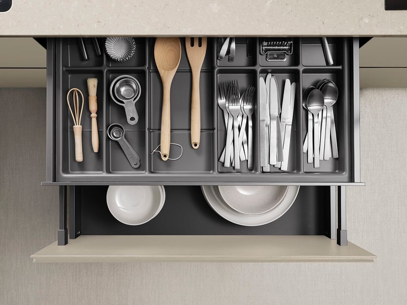 Besticklåda med organiserade köksredskap, inklusive bestick, träredskap, visp, och måttskedar, samt två tallrikar i en låda under