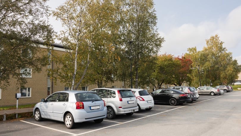 Parkeringsplats med flera bilar, omgiven av träd och en byggnad i bakgrunden