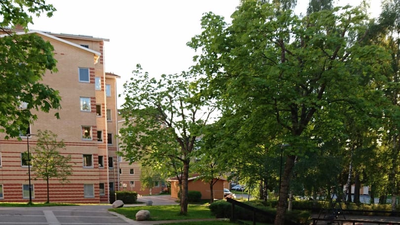 Bostadsområde med flerfamiljshus byggda i ljus tegel, omgivna av gröna träd och en gångväg