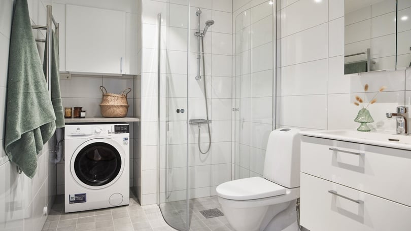 Modernt badrum med vit kakelvägg, glasdusch, tvättmaskin, vitt skåp och toalett