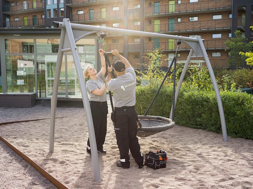 Två personer i arbetskläder installerar eller reparerar en gungställning med en korggunga på en lekplats, omgiven av moderna byggnader och grönska