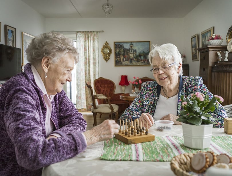 Två äldre kvinnor spelar brädspel vid ett bord i ett vardagsrum
