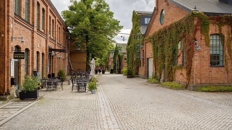 Tom gata med kullersten mellan röda tegelbyggnader, uteservering och grönskande fasader