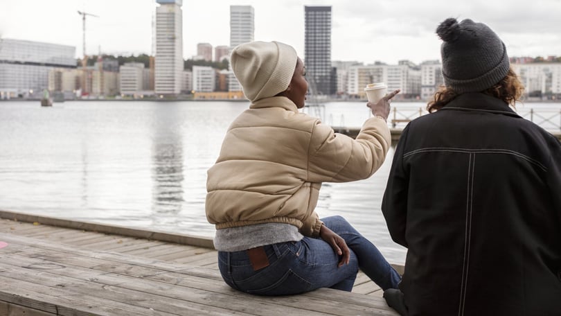 Två personer sitter vid vattnet och tittar ut mot stadens skyline, varav en håller en kopp