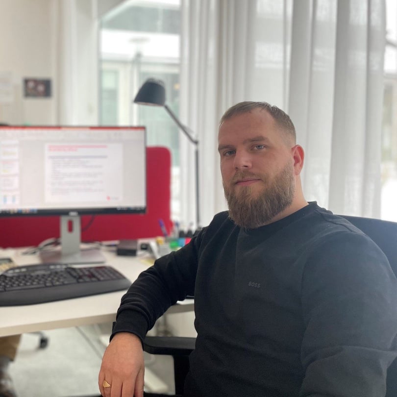 Porträtt av en man i svart tröja som sitter vid ett skrivbord med datorskärm och tangentbord