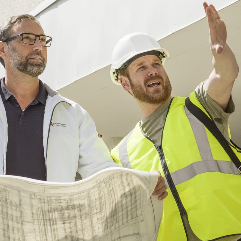 Två byggarbetare tittar på en ritning, varav den ena bär hjälm och varselväst