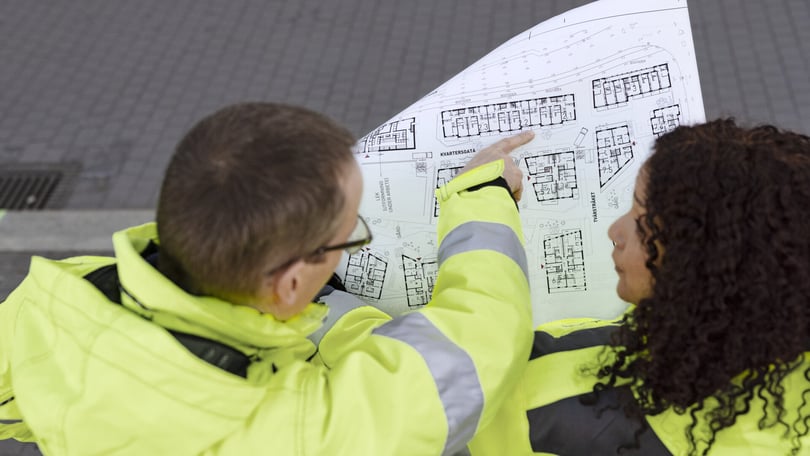 Två personer i gula reflexjackor studerar en ritning med bostadslayouter
