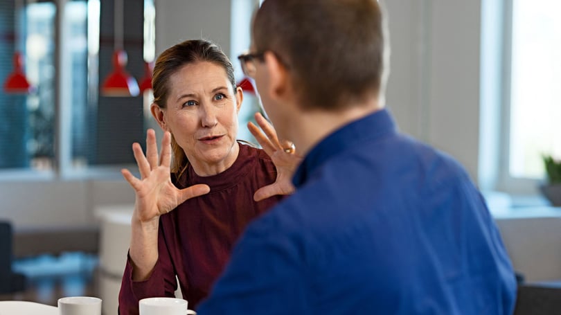 Två personer i samtal inomhus, där en kvinna gestikulerar med händerna och en man ses i förgrunden