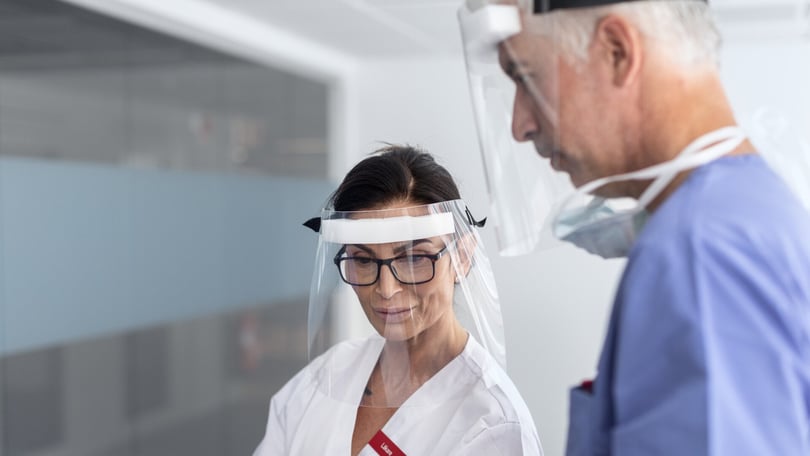 Två sjukvårdspersonal i munskydd och visir, en kvinna i vit vårdklädsel och en man i blå operationskläder