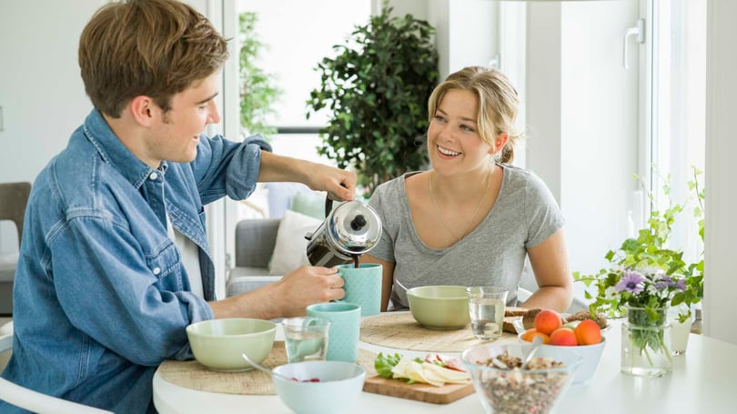 Två unga personer vid ett köksbord, en man häller kaffe i en kopp