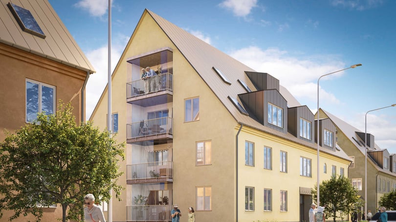 Gult flerfamiljshus med balkonger, omgiven av träd och en trottoar under en klarblå himmel