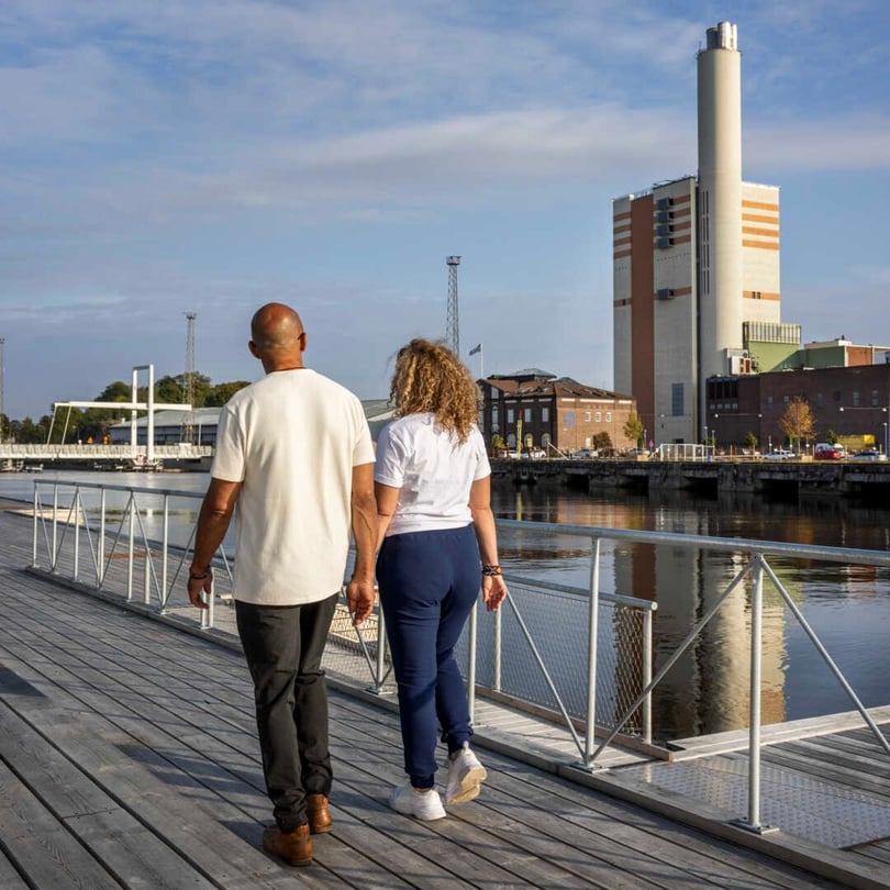 Två personer promenerar längs en träbrygga vid vattnet i Norrköping med en industribyggnad i bakgrunden