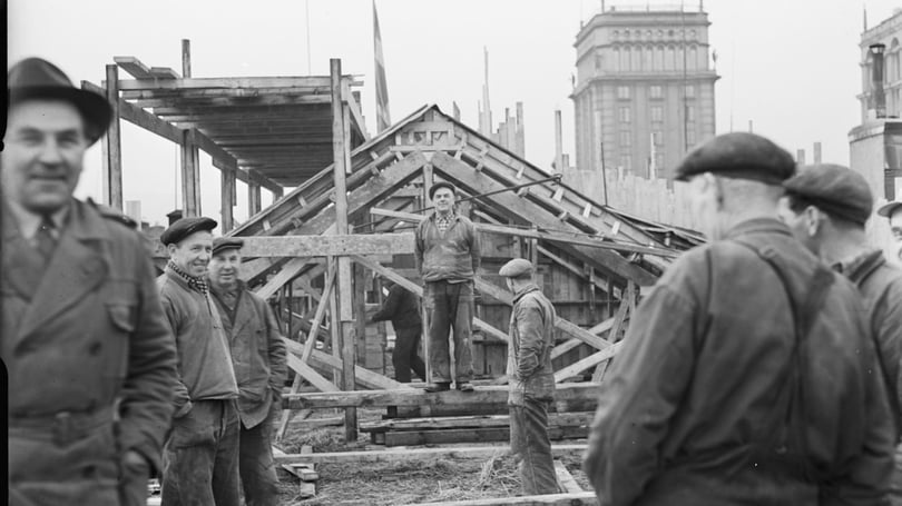 Svartvit bild av flera byggarbetare vid en huskonstruktion, med trästomme och stadshus i bakgrunden