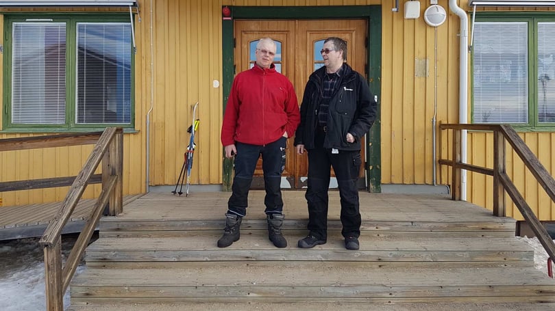 Två personer står på en träveranda framför en gul byggnad