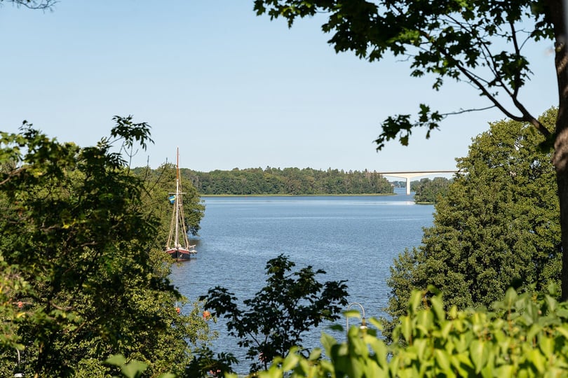 Vy över en sjö med segelbåt, grönska i förgrunden och en bro i bakgrunden