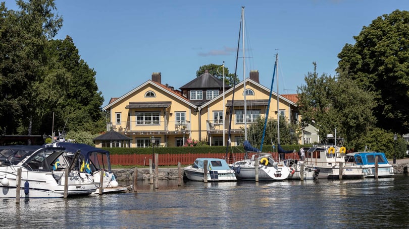 Gul sekelskiftesvilla i Trosa vid vattnet med förtöjda båtar i förgrunden och grönskande träd runtom