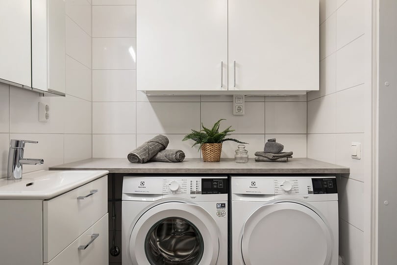 Tvättstuga med tvättmaskin och torktumlare under en bänkskiva, vitt kaklade väggar och handfat