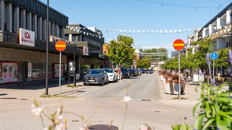 Gågata med butiker, parkerade bilar och blomsterplanteringar i ett bostadsområde