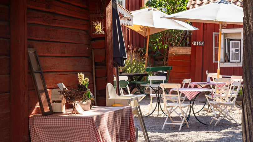 Uteservering med bord, stolar och vita parasoller framför röda träbyggnader