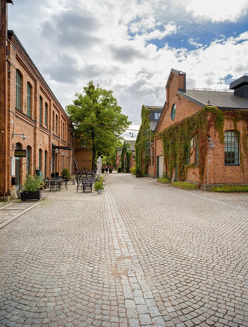 Tom gata mellan röda tegelbyggnader med uteservering, gröna växter och molnig himmel
