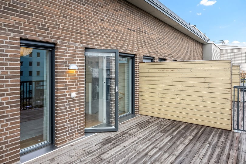 Balkong med trätrall, tegelvägg, glasdörrar och träpanel, omgiven av byggnader under klarblå himmel
