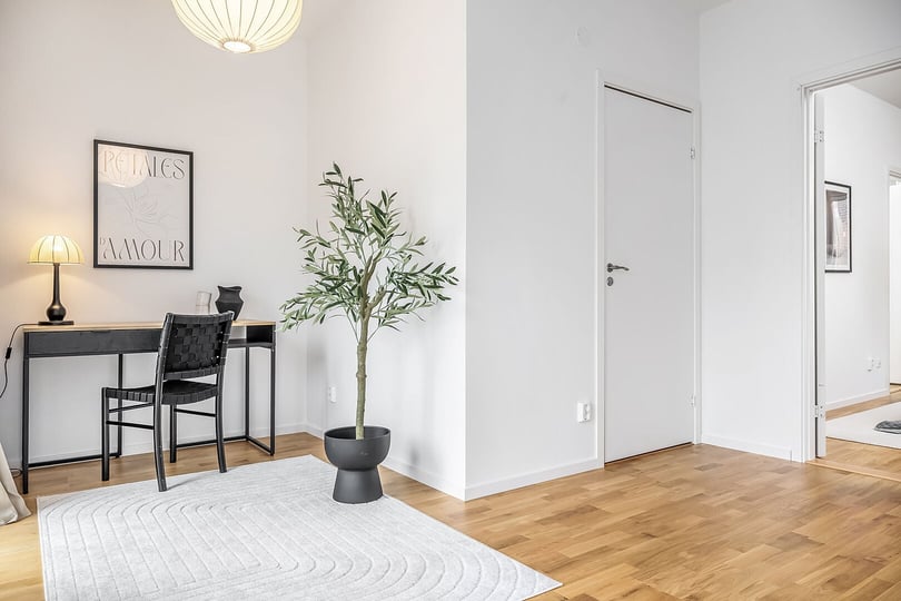 Ljus inredning med ett skrivbord, stol och bordslampa bredvid en växt i kruka på ett vitt matta. Väggarna är vita, och dörrar leder till olika rum
